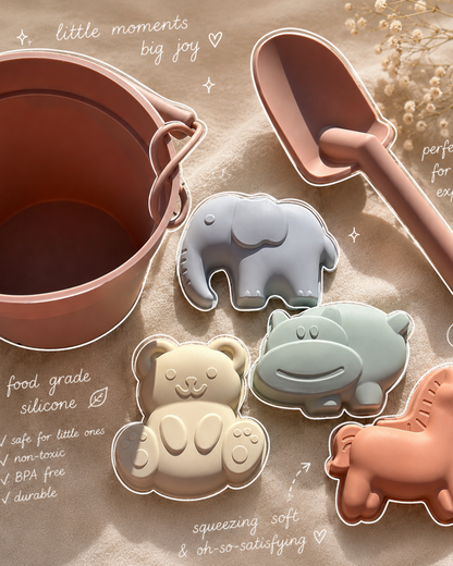 A children's silicone mold set arranged on a sandy surface, including a pinkish-brown bucket, a matching scoop, and four animal-shaped molds: an elephant, hippo, bear, and horse. Handwritten text overlays describe the product benefits.